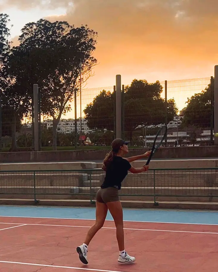 Nicole finishes a stroke on a hard court at sunset, under an orange sky.