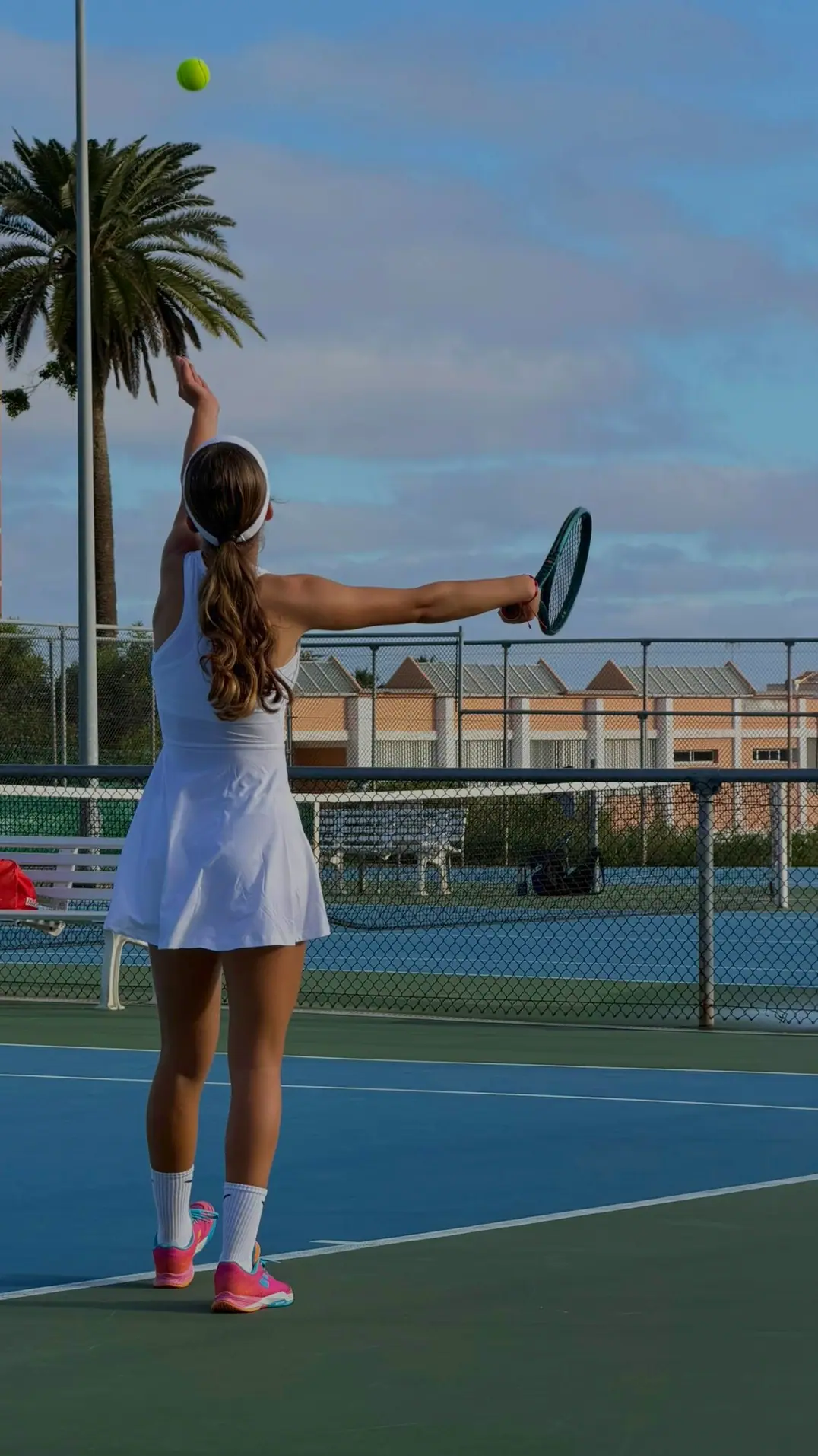 Nicole, seen from behind in a white dress, tosses the ball to serve on a blue court; behind her, a palm tree and a cloudy sky.