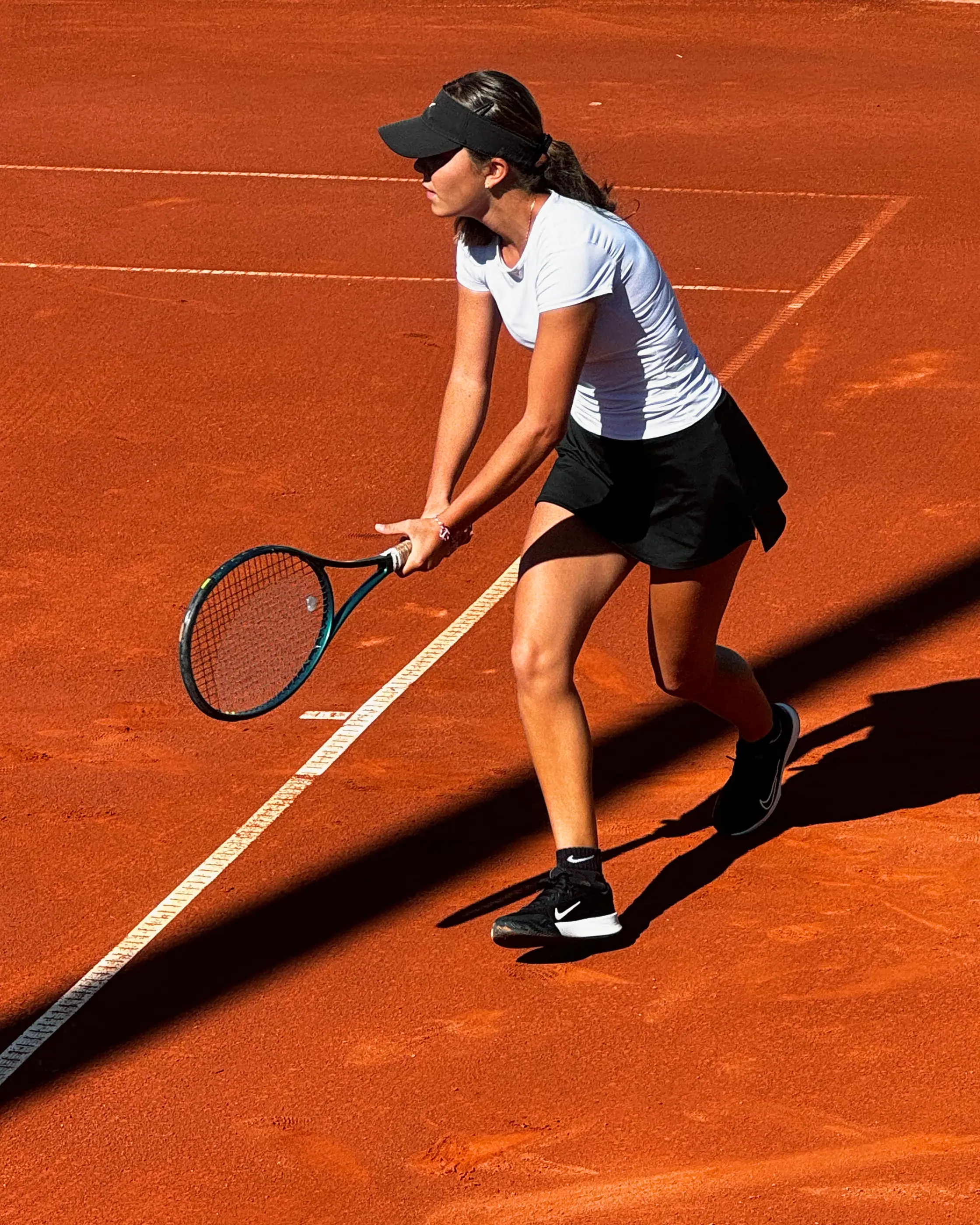 Nicole in a ready stance on a clay court, seen from above: knees bent, racket held low in both hands, a long shadow stretching across the court.