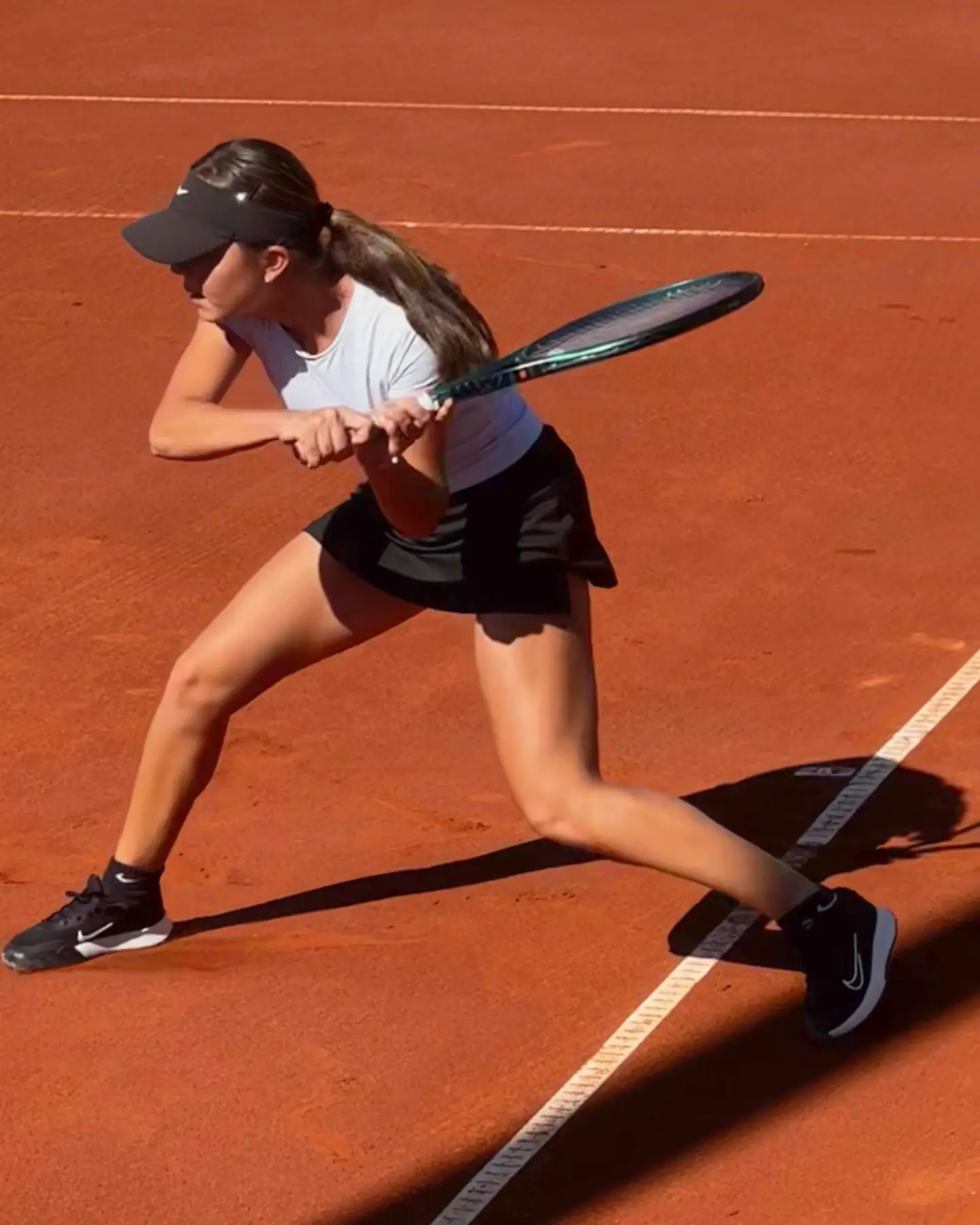 Nicole stepping into a two-handed backhand on clay, seen from above, racket taken back and a long shadow across the court.