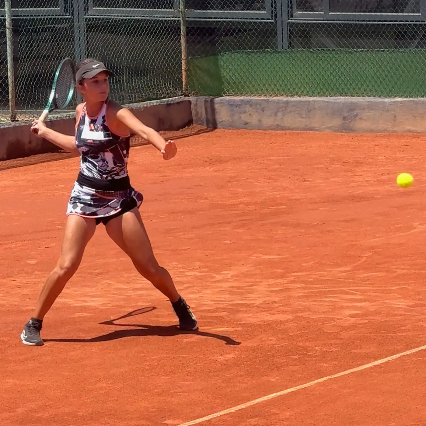 Nicole facing the camera in a wide stance on a clay court, racket drawn back for a forehand, with the ball in the air to one side.
